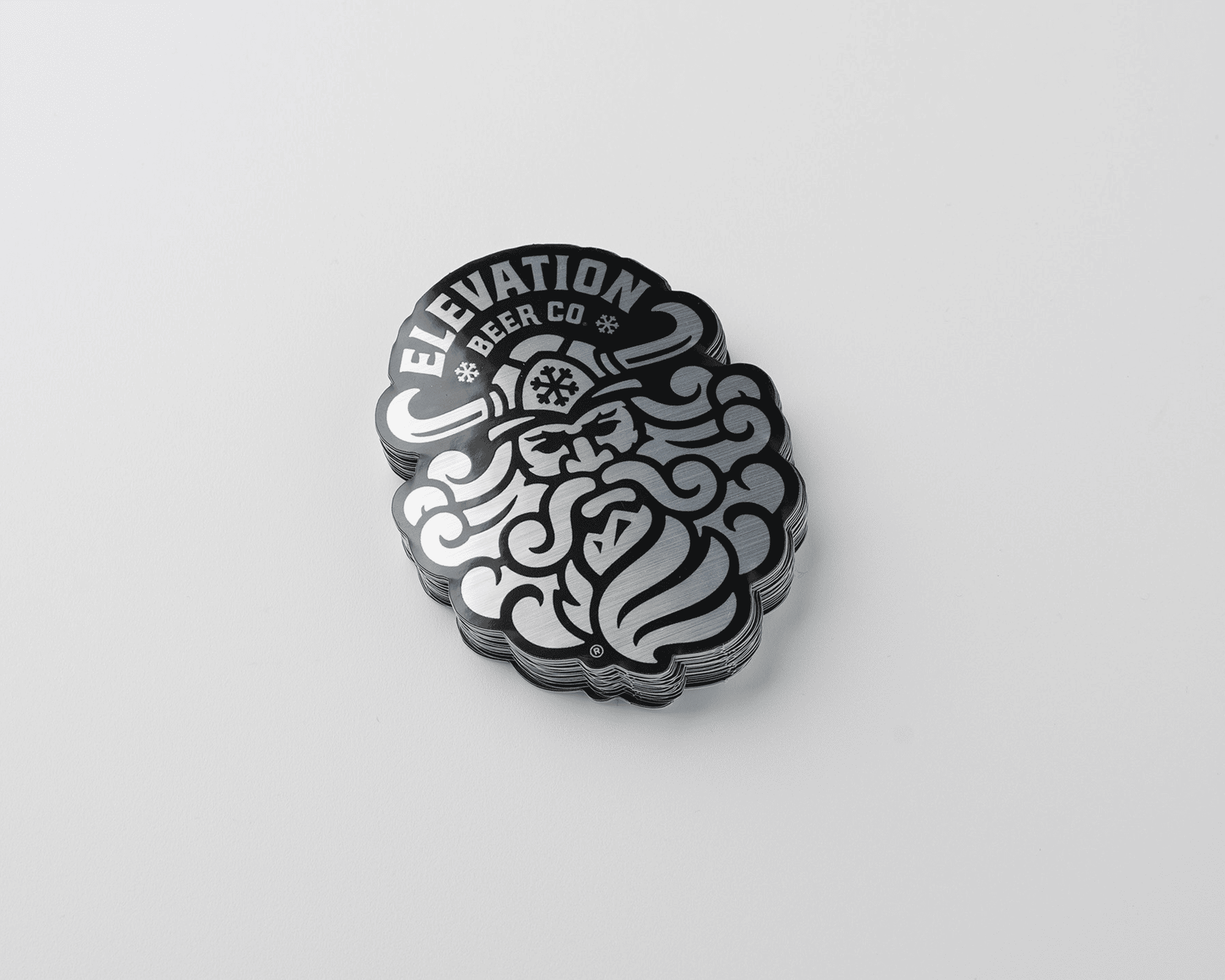 Stack of black and silver brushed aluminium stickers featuring a stylized, intricate design of a bearded face with "Elevation Beer Co." text.